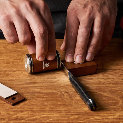 Hands use the Tumbler Mini Knife Sharpener on a wooden table with the kit laid out nearby